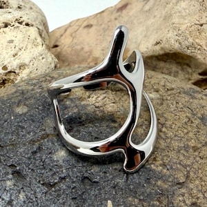 May include: A polished silver-toned ring featuring an open, asymmetrical design with smooth, curved lines that create a branching, organic shape. The metal surface is highly reflective, showcasing a sleek, continuous form without any gemstones.
