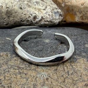 May include: A polished silver-toned metal ring featuring an open, irregular, and wavy band design. The ring has a smooth, reflective surface and a gap between the two ends, creating a continuous, fluid, and organic shape that rests on a stone.