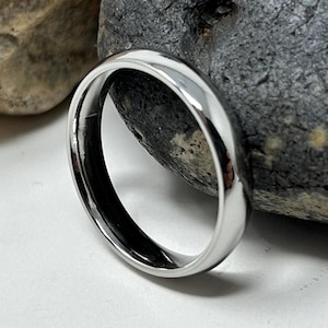 May include: A polished silver ring, reflecting light, set against a backdrop of two stones, one light brown and the other dark grey. The ring's smooth surface highlights its simple design. A piece of jewellery.