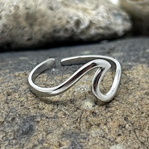 May include: A silver ring with a wave design. The ring is open at the back and rests on a grey stone. The background features other grey and beige stones. This ring is a piece of jewellery.