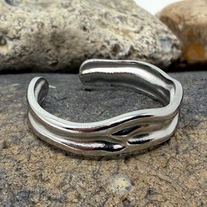 May include: A silver-toned open ring featuring a smooth, undulating, and organic wave-like design. The metal band has a polished, reflective surface with a continuous, flowing shape that creates a gap at the top, allowing for adjustable sizing.