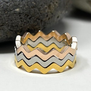 Könnte beinhalten: Drei übereinanderliegende Ringe mit einem wellenförmigen Design. Die Ringe sind in Gold, Silber und Roségold.