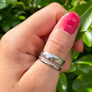 May include: A silver ring with a double-band design. The ring is worn on a finger, with bright pink nail polish visible. The background features green leaves.