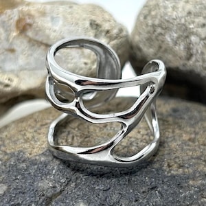 Op de afbeelding: Een zilverkleurige ring met een open, abstract ontwerp. De ring heeft meerdere onderling verbonden banden met onregelmatige uitsparingen. De ring wordt getoond op een donkergrijze steen, met andere stenen op de achtergrond.