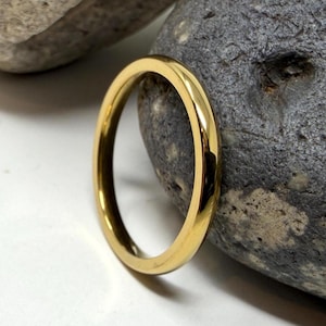 May include: A gold ring with a polished finish. The ring is circular and rests on a white surface beside two grey stones. The ring is a simple band, suitable for daily wear or as a wedding ring. The ring's diameter is approximately 2 cm.
