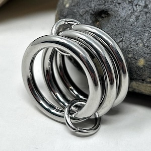 Puede incluir: Un anillo de metal plateado brillante con múltiples bandas circulares conectadas. Los anillos están apilados, con un anillo más pequeño adjunto en la parte inferior. El objeto está sobre un fondo blanco con una roca.