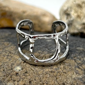 May include: A silver ring with a geometric design. The ring is made of metal and has a hammered texture. The ring is sitting on a rock.
