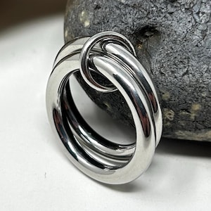Puede incluir: Un anillo plateado brillante con un diseño único. El anillo presenta tres bandas conectadas, creando un efecto en capas. El metal tiene un acabado pulido, reflejando la luz. El anillo está sobre un fondo neutro con una piedra.