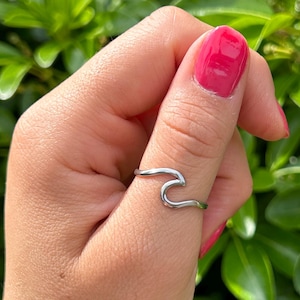 May include: A silver-colored ring in the shape of a wave. The ring is worn on a finger, with a background of green leaves. The ring's design is simple and elegant, with a smooth, curved shape.