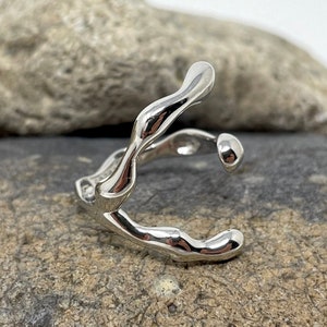 Puede incluir: Un anillo plateado con un dise&ntilde;o org&aacute;nico y fluido. El anillo, probablemente de plata, tiene una forma &uacute;nica y abstracta. Se muestra sobre una superficie rocosa gris. El dise&ntilde;o recuerda al agua que fluye o al metal fundido.