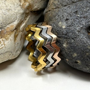 Könnte beinhalten: Drei übereinanderliegende Ringe mit einem Zickzackmuster. Die Ringe sind in Gold, Silber und Roségold.