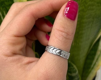 May include: A silver ring with a repeating chevron pattern. The ring is worn on a finger, with a green and yellow plant in the background. The ring's design is a series of angled lines, creating a textured appearance.