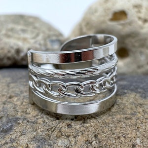 May include: A silver ring with a layered design featuring a chain, rope, and two flat bands.