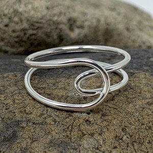 Può includere: Un anello d'argento con un designo semplice e moderno. L'anello è composto da due bande di filo, una delle quali è attorcigliata a forma di cerchio.