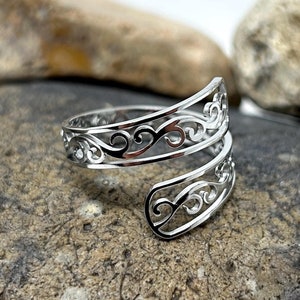 May include: A silver ring featuring an open filigree design with swirling patterns. The ring has an adjustable, wrap-around style. It is shown on a grey and brown stone surface.
