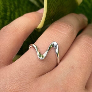 Könnte beinhalten: Ein silberner Ring mit einem wellenförmigen, abstrakten Design. Der Ring wird an einem Finger getragen und der Hintergrund ist eine grüne Pflanze.