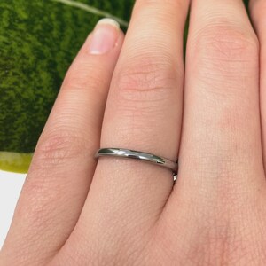 May include: A silver ring is worn on a finger, with green and yellow plant leaves in the background. The ring is thin and metallic. The image focuses on the ring and the hand, with the plant providing a natural backdrop.
