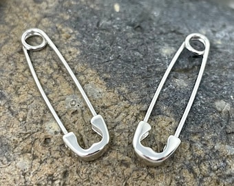 May include: Two silver safety pin earrings. The earrings are on a gray textured surface.