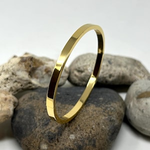 Könnte beinhalten: Ein goldfarbenes Armreif-Armband mit glatter, polierter Oberfläche. Das Armband ist aus Metall und hat ein schlichtes, elegantes Design.