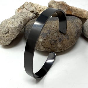 May include: A smooth, matte black cuff bracelet with a simple, open-ended design. The bracelet features a solid, uniform band with a dark, non-reflective finish that curves into a circular shape, showcasing a clean and straightforward aesthetic.