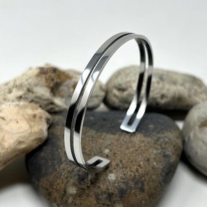 May include: A silver-toned metal cuff bracelet featuring a double-band design with a smooth, polished finish. The bracelet has an open-ended structure with rectangular terminals at each end, creating a clean and reflective appearance.
