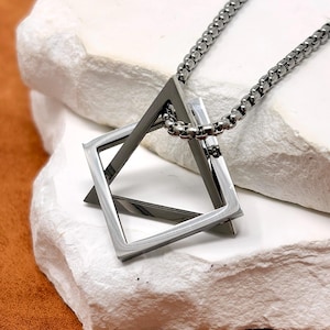 May include: A silver-coloured geometric pendant necklace with a square and a triangle design. The pendant hangs from a matching chain. The necklace is displayed on a white, textured surface.