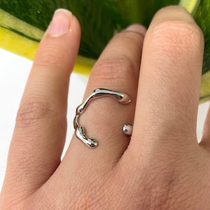 Könnte beinhalten: Ein silberfarbener Ring mit organischem, fließendem Design. Der Ring scheint aus glänzendem Metall zu bestehen und hat eine einzigartige, abstrakte Form. Der Ring wird am Finger getragen.