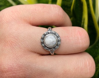 May include: A silver ring with a white moonstone set in a circular bezel with a beaded border.