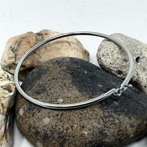 May include: A silver bangle bracelet with a distinctive clasp. The circular bracelet is displayed on a collection of smooth, grey and tan stones. The clasp has a decorative, textured design.