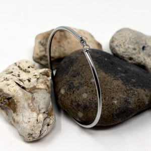 May include: A thin, polished silver-tone metal bangle bracelet with a smooth, circular band and a small, integrated clasp mechanism at the top. The bracelet features a reflective, metallic finish and a simple, continuous design.