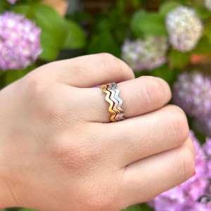May include: A stack of three thin metal rings worn on a finger. Each ring features a distinct zigzag or chevron pattern. The set includes one gold-toned ring, one rose gold-toned ring, and one silver-toned ring, all worn together as a single band.