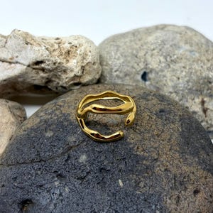Puede incluir: Un anillo dorado con un dise&ntilde;o ondulado y org&aacute;nico. El anillo abierto se encuentra sobre una roca gris oscura, rodeado de otras rocas. La superficie del anillo refleja la luz, resaltando su textura suave y color dorado.