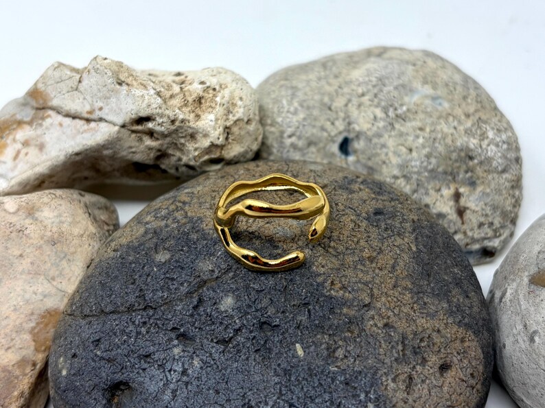 Puede incluir: Un anillo dorado con un dise&ntilde;o ondulado y org&aacute;nico. El anillo abierto se encuentra sobre una roca gris oscura, rodeado de otras rocas. La superficie del anillo refleja la luz, resaltando su textura suave y color dorado.