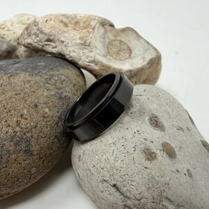 May include: A black ring with a flat top and a beveled edge rests on a light-colored stone. The ring is made of a dark, shiny material. The background features other stones.