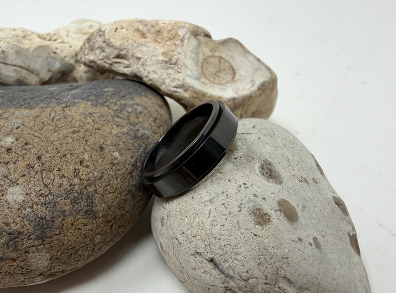 May include: A black ring with a flat top and a beveled edge rests on a light-colored stone. The ring is made of a dark, shiny material. The background features other stones.