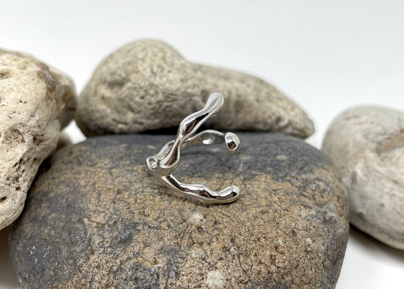 Puede incluir: Un anillo plateado con un dise&ntilde;o org&aacute;nico y fluido. El anillo, probablemente de plata, tiene una forma &uacute;nica y abstracta. Se muestra sobre una superficie rocosa gris. El dise&ntilde;o recuerda al agua que fluye o al metal fundido.