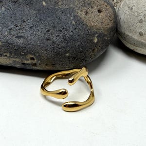 Puede incluir: Un anillo dorado con un dise&ntilde;o abstracto y fluido. El anillo tiene una forma &uacute;nica con extremos redondeados y una abertura central. Se encuentra sobre una superficie blanca, con dos piedras en el fondo.