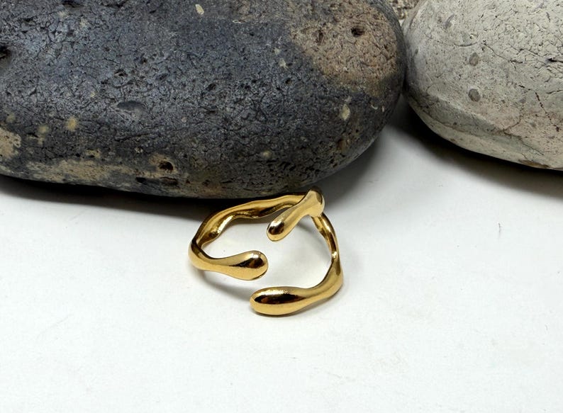 Puede incluir: Un anillo dorado con un dise&ntilde;o abstracto y fluido. El anillo tiene una forma &uacute;nica con extremos redondeados y una abertura central. Se encuentra sobre una superficie blanca, con dos piedras en el fondo.
