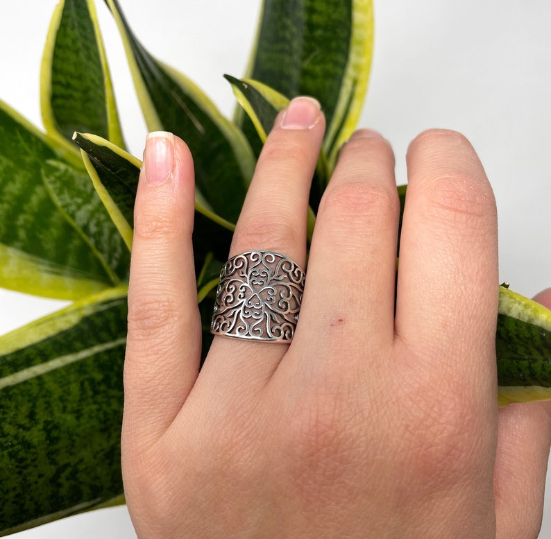 May include: A silver filigree ring with a floral design. The ring is wide and has a delicate, intricate pattern.