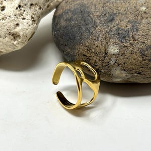 May include: A gold-colored adjustable ring with an abstract design. The ring has an open design with a textured surface and is set against a white background with rocks.