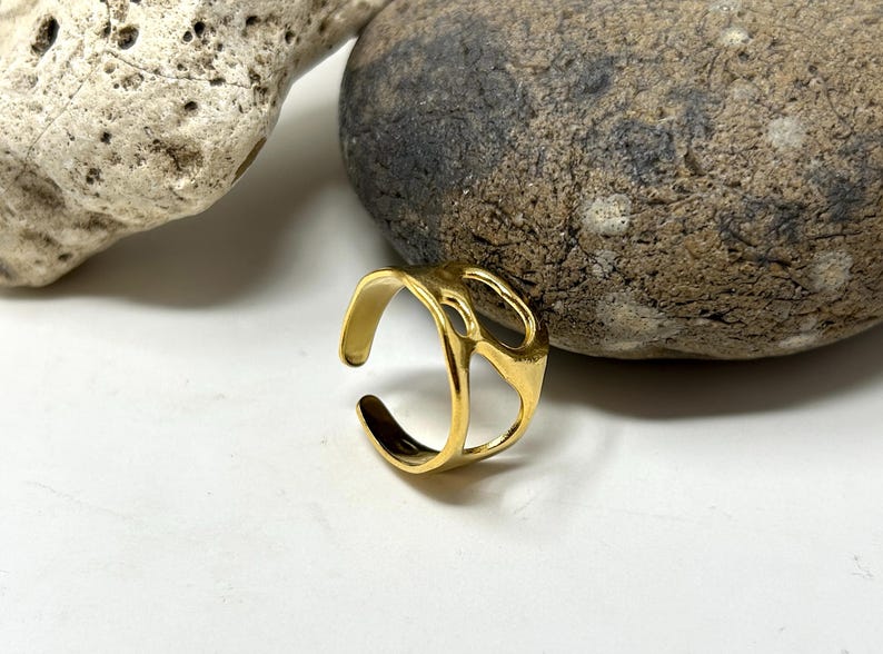 May include: A gold-colored adjustable ring with an abstract design. The ring has an open design with a textured surface and is set against a white background with rocks.