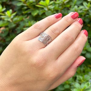 May include: A silver ring with a delicate, openwork design resembling a tree branch. The ring is worn on a finger with pink nail polish.