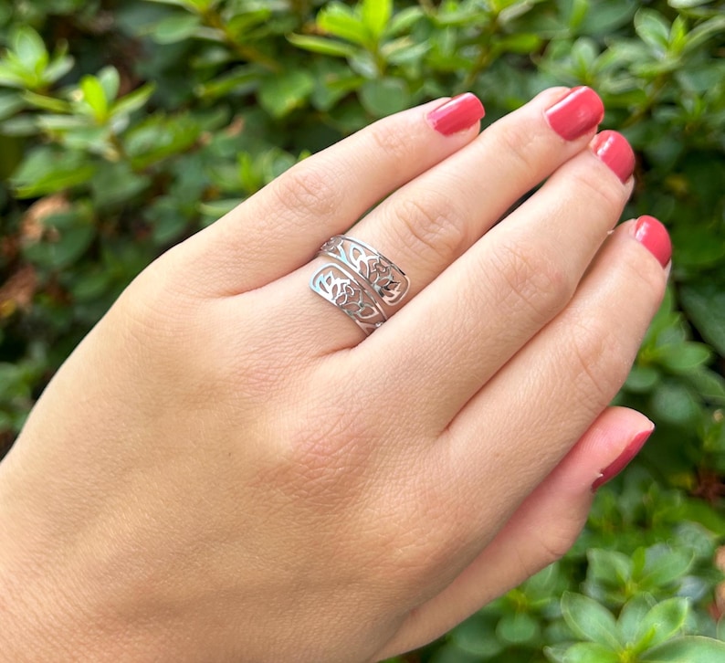 May include: A silver ring with a delicate, openwork design resembling a tree branch. The ring is worn on a finger with pink nail polish.