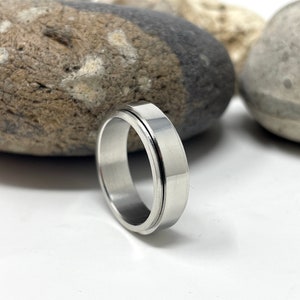 May include: A silver-colored ring with a flat, wide band and a grooved outer edge. The ring is made of a shiny, metallic material and is displayed on a white surface with blurred rocks in the background. The ring is a piece of jewelry.