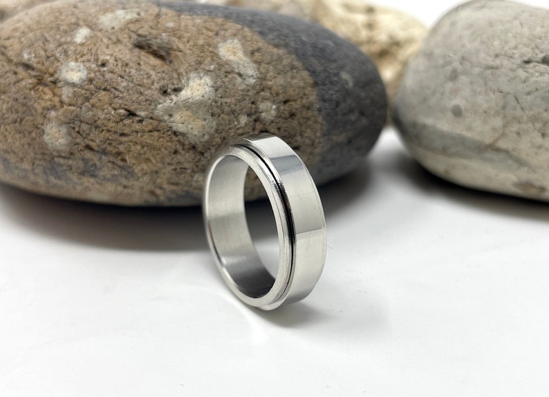 May include: A silver-colored ring with a flat, wide band and a grooved outer edge. The ring is made of a shiny, metallic material and is displayed on a white surface with blurred rocks in the background. The ring is a piece of jewelry.