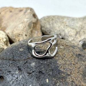 May include: A silver-colored adjustable ring with an abstract, openwork design. The ring features irregular, organic shapes and is displayed on a dark gray rock. The ring's surface reflects light, highlighting its metallic finish.