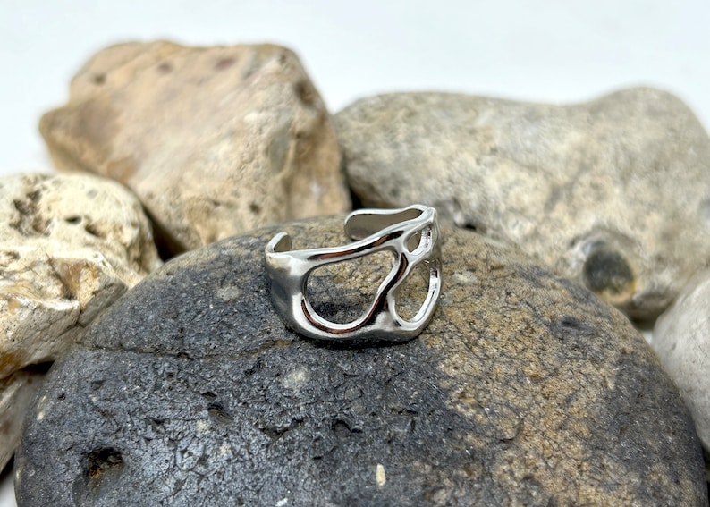 May include: A silver-colored adjustable ring with an abstract, openwork design. The ring features irregular, organic shapes and is displayed on a dark gray rock. The ring's surface reflects light, highlighting its metallic finish.