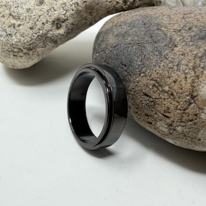 May include: A black ring with a matte finish sits on a white surface next to a large, textured rock. The ring is circular and appears to be made of metal. The ring's design is simple and modern.