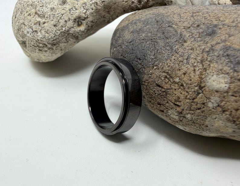 May include: A black ring with a matte finish sits on a white surface next to a large, textured rock. The ring is circular and appears to be made of metal. The ring's design is simple and modern.