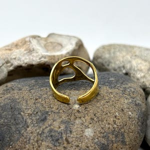 May include: A gold-colored adjustable ring with an open design. The ring is set against a backdrop of gray and tan stones. The ring's design features an open, abstract shape.
